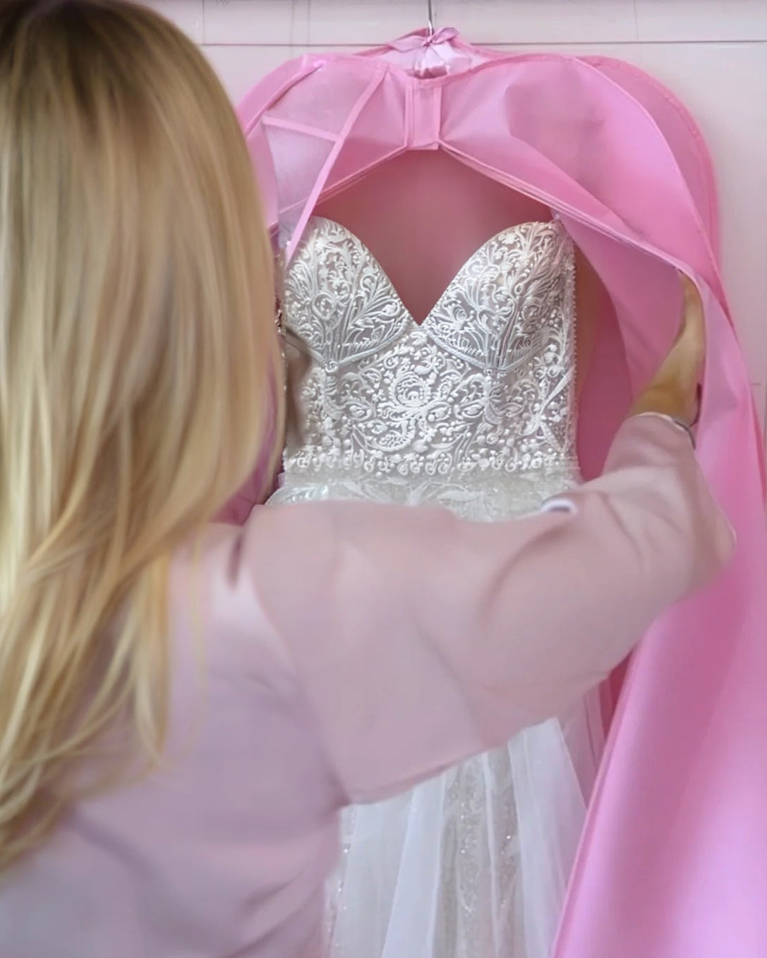 A woman in a light pink robe is seen from behind, lifting a bright pink bridal veil or dress cover to reveal a white wedding gown hanging on a gold hanger against a pink wall.